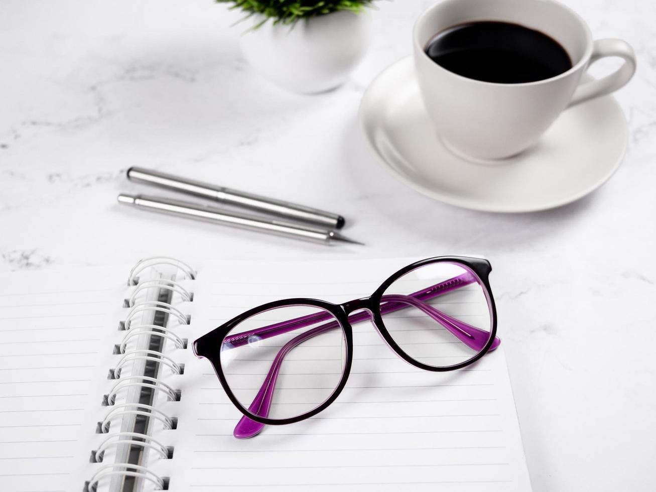 gafas de primer plano en un cuaderno de página en blanco con bolígrafo plateado y taza de café en el fondo de mármol foto