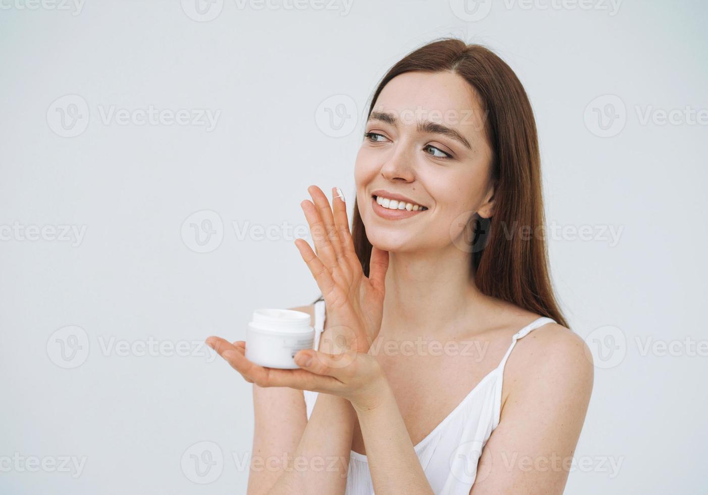 retrato de belleza de una mujer feliz y sonriente con el pelo largo y oscuro puesto crema hidratante nutritiva de día en la cara y las manos limpias y frescas de la piel en el fondo blanco aislado foto