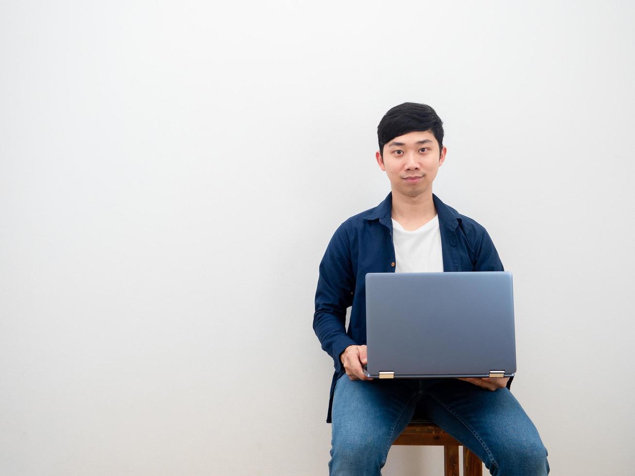 hombre asiático hermosa sonrisa feliz sentado en la silla con la computadora portátil en el fondo de la pared blanca foto