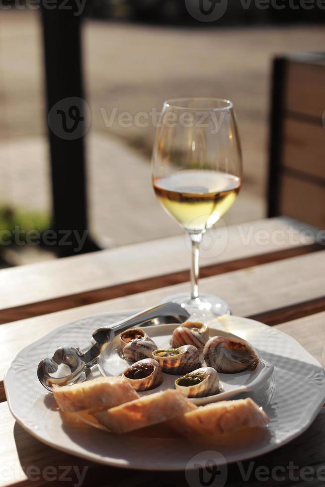 vaso con vino y plato con las conchas de caracol, servido en un plato clásico blanco, con varias salsas, pesto, trufa, queso, trozos de baguette francés, tenedor para una concha, mesa de madera en café al aire libre foto