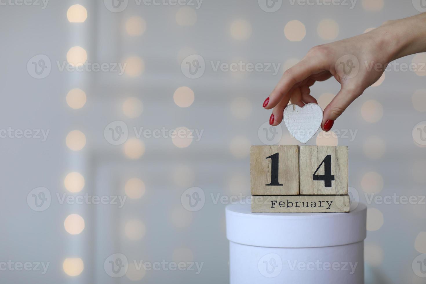 Valentine's day concept. Date 14 February on wooden cube calendar on a white gift box and white heart in hand. Bokeh golden and white background. photo