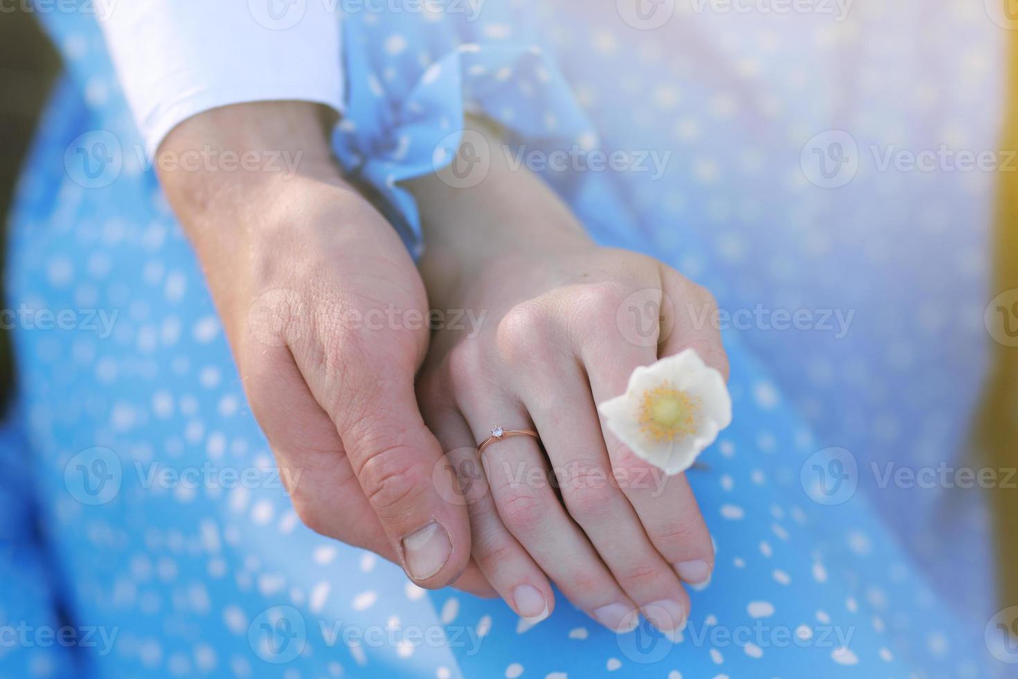 concepto de cita romántica. manos de hombre y mujer jóvenes abrazándose unos a otros. chica con anillo de oro en el dedo en vestido azul con flor de manzanilla en la mano foto