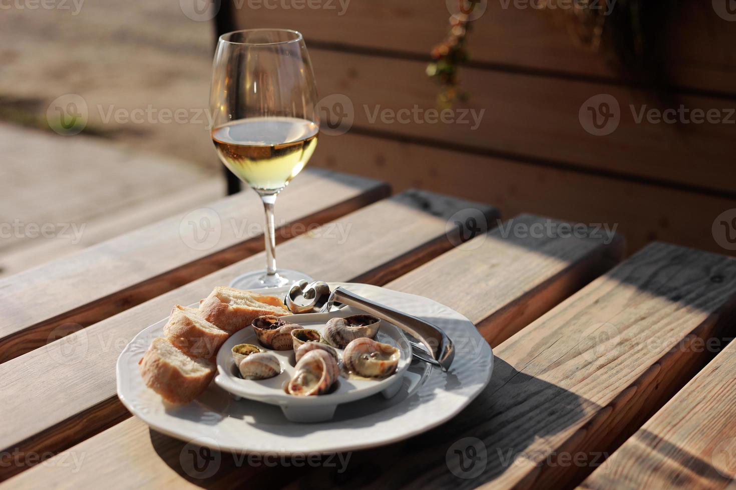 Glass with wine and dish with the snail shells, served on a white classic plate, with various sauces, pesto, truffle, cheese, pieces of French baguette, fork for a shell, wooden table in outdoor cafe photo