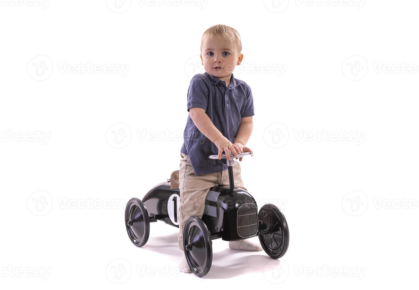 niño montando un coche de juguete de estilo retro. niño montando un viejo coche de pedales de metal para niños del siglo XIX. aislado sobre fondo blanco foto