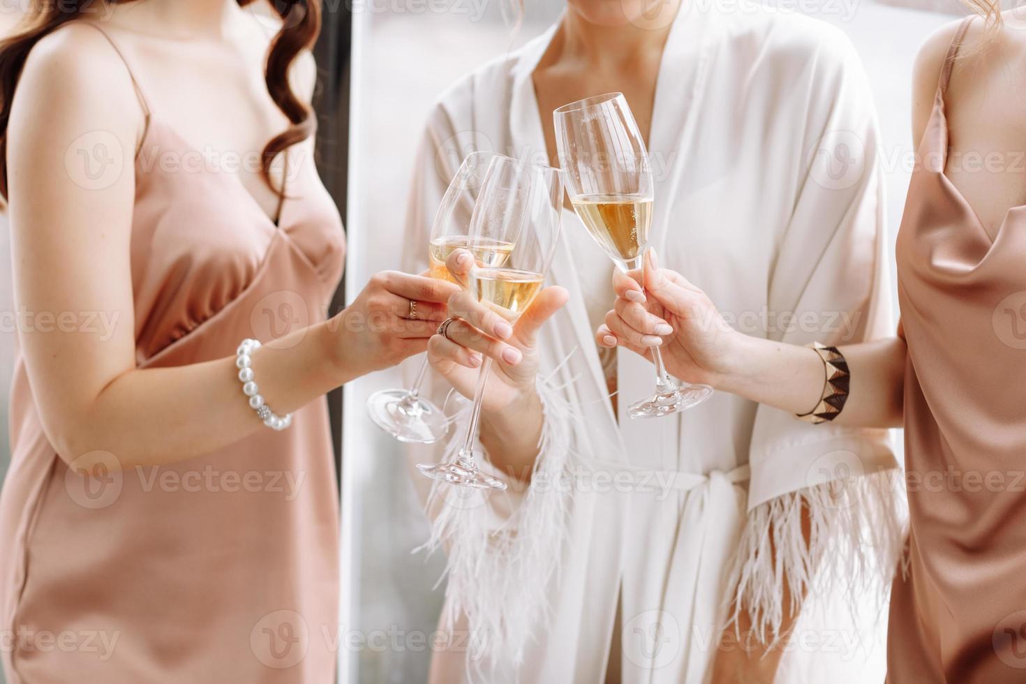 Morning of bride. Gorgeous bride with best bridesmaids are holding glasses and drinking champagne in hotelroom near the large window. Sexy bridesmaids in exciting negligee. Wedding morning details photo