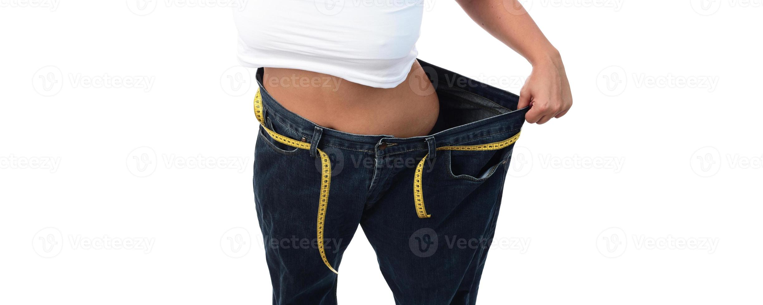 mid section of a woman with belly fat holding her blue jeans. Girl with big jeans isolated on white background. Woman shows her weight loss. Healthy lifestyle concept photo