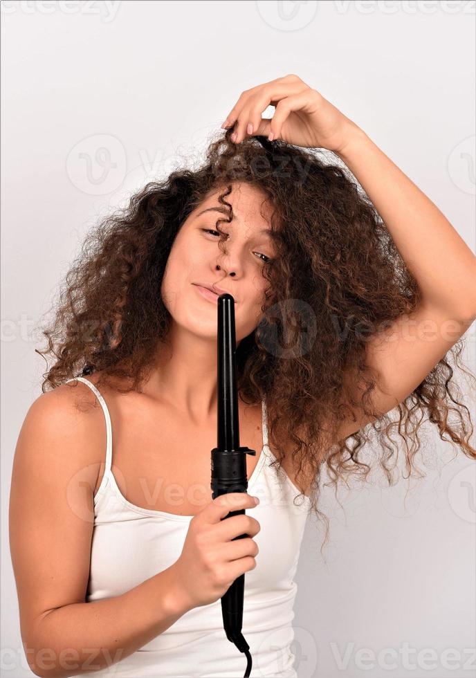 Pelo RIZADO. bella mujer sonriente con el pelo largo y ondulado planchándolo, usando un rizador. chica feliz con un hermoso cabello suave y saludable usando un rizador para rizos perfectos. peinado y peluqueria foto