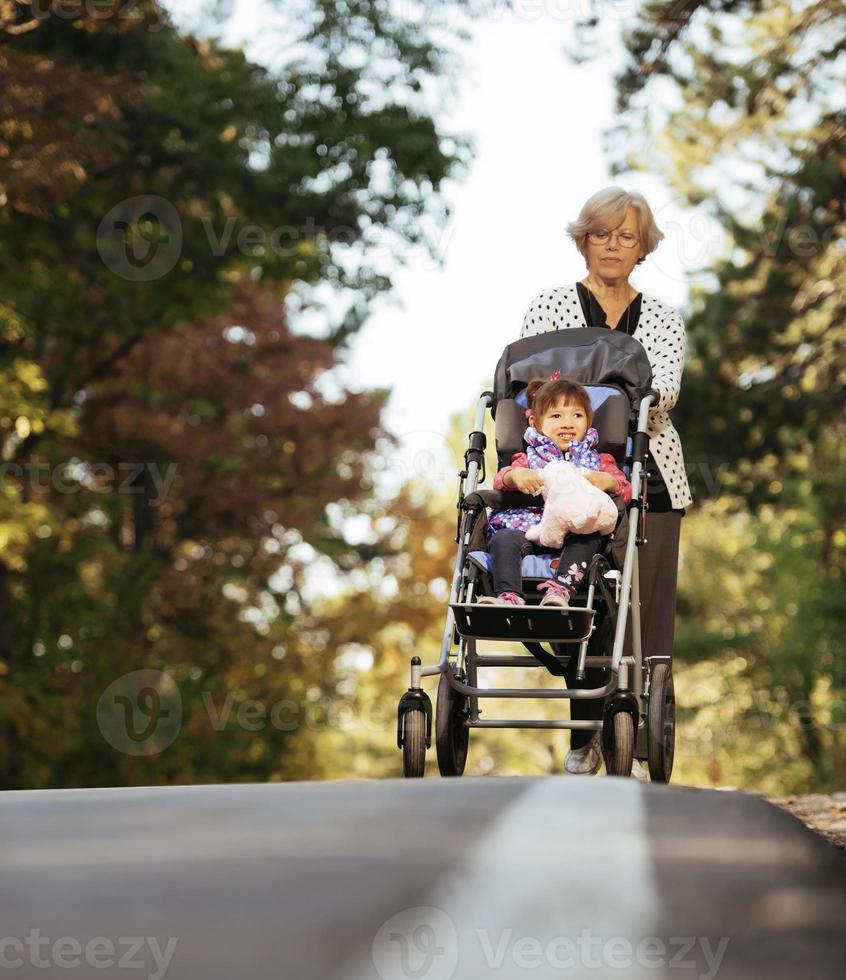 Happy senior lady pushing wheel chair and children. Grandmother and kids enjoying a walk in the park. Child supporting disabled grandparent. Family visit. Generations love and relationship photo