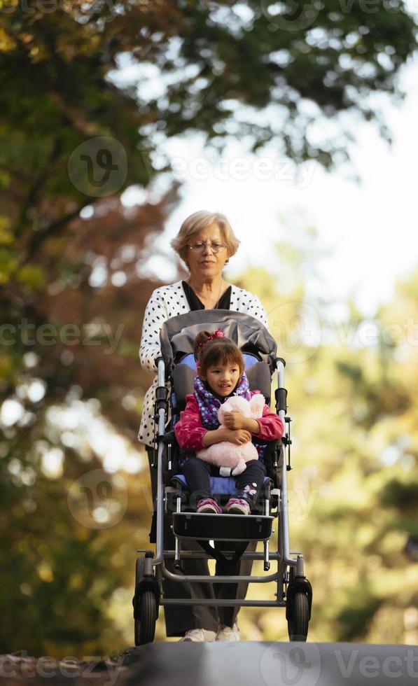 Happy senior lady pushing wheel chair and children. Grandmother and kids enjoying a walk in the park. Child supporting disabled grandparent. Family visit. Generations love and relationship photo