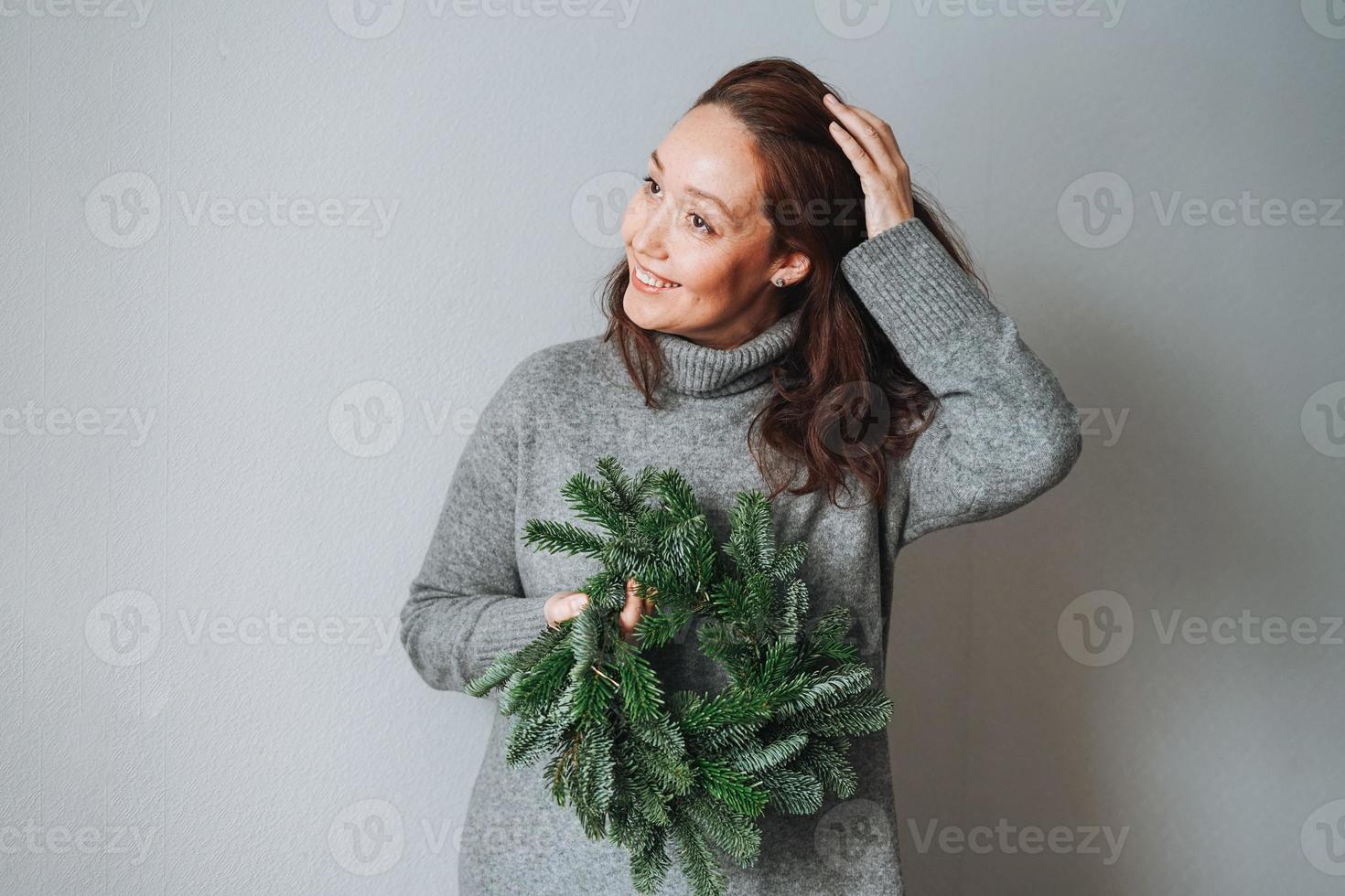 Adult beautiful smiling woman forty years with brunette curly hair in warm grey knitted dress with diy fir christmas wreath in hand on the grey background photo