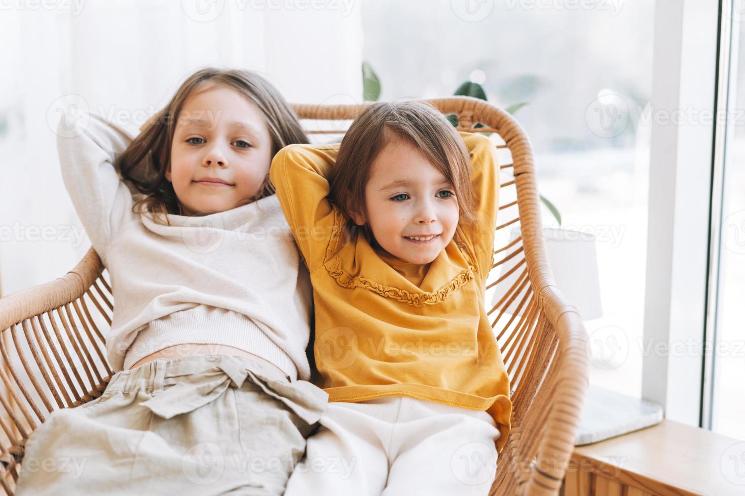 Cute little sisters girls on a wicker chair near window at the home photo