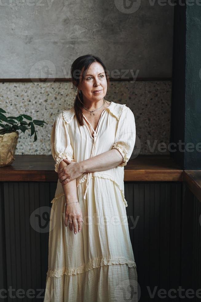 Portrait of friendly adult middle aged brunette smiling woman fifty years in white dress in kitchen with green house plants at home photo