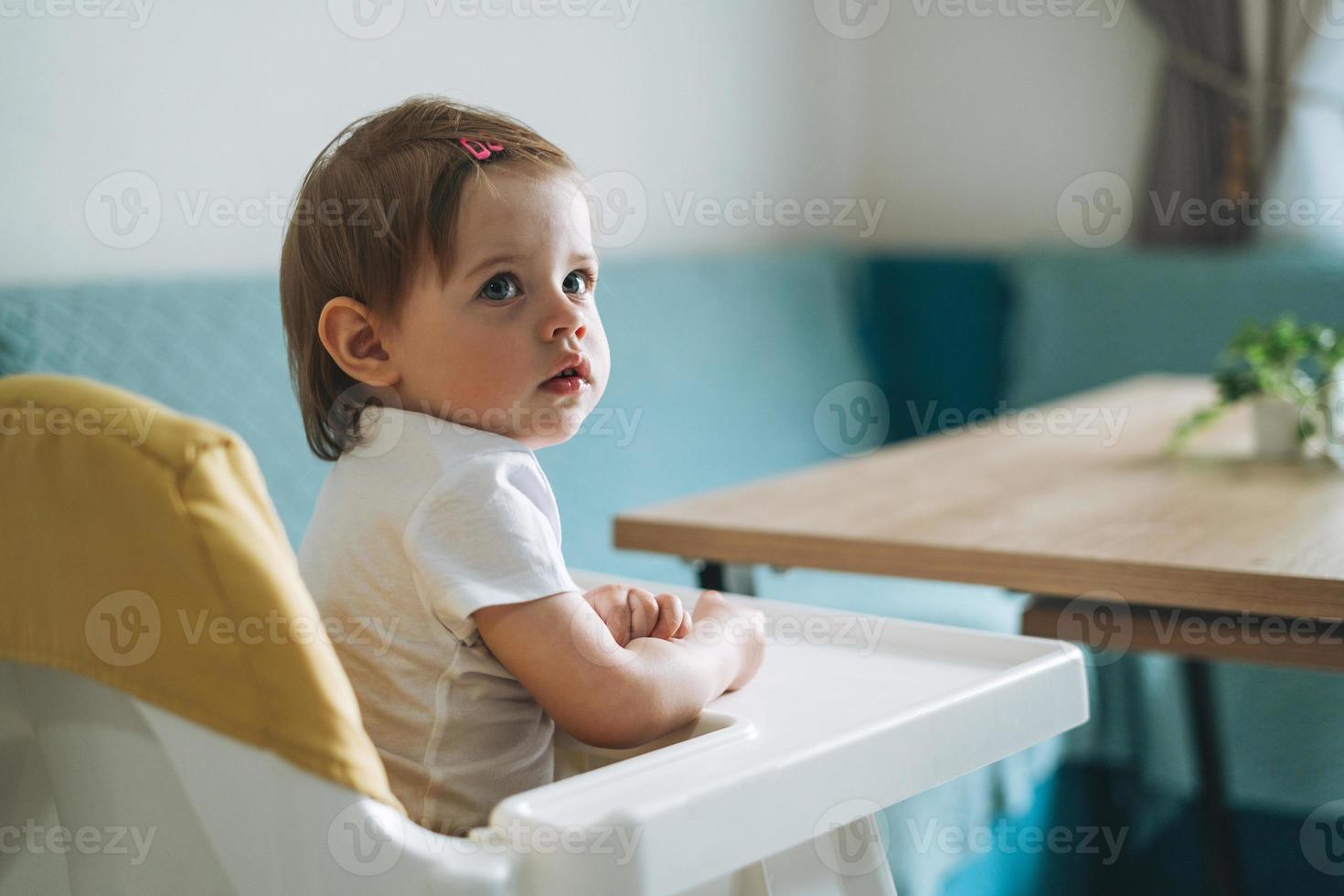 Charming little baby girl sitting in high chair for feeding at the bright kitchen at home photo