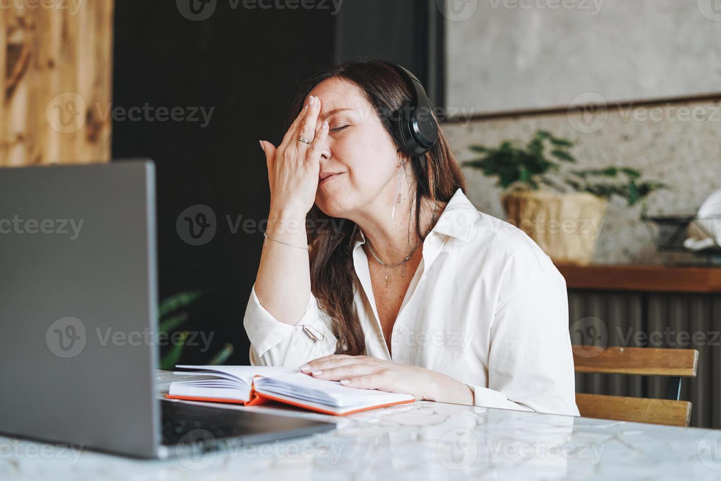 Adult tired brunette woman fifty years in headphones doing notes in daily book with opened laptop,  study something with online course at the home photo