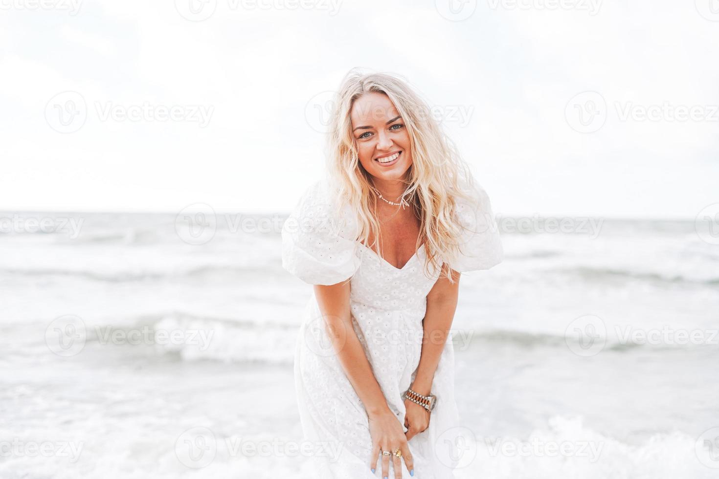 joven mujer rubia hermosa con el pelo largo en vestido blanco disfrutando de la vida en la playa del mar foto