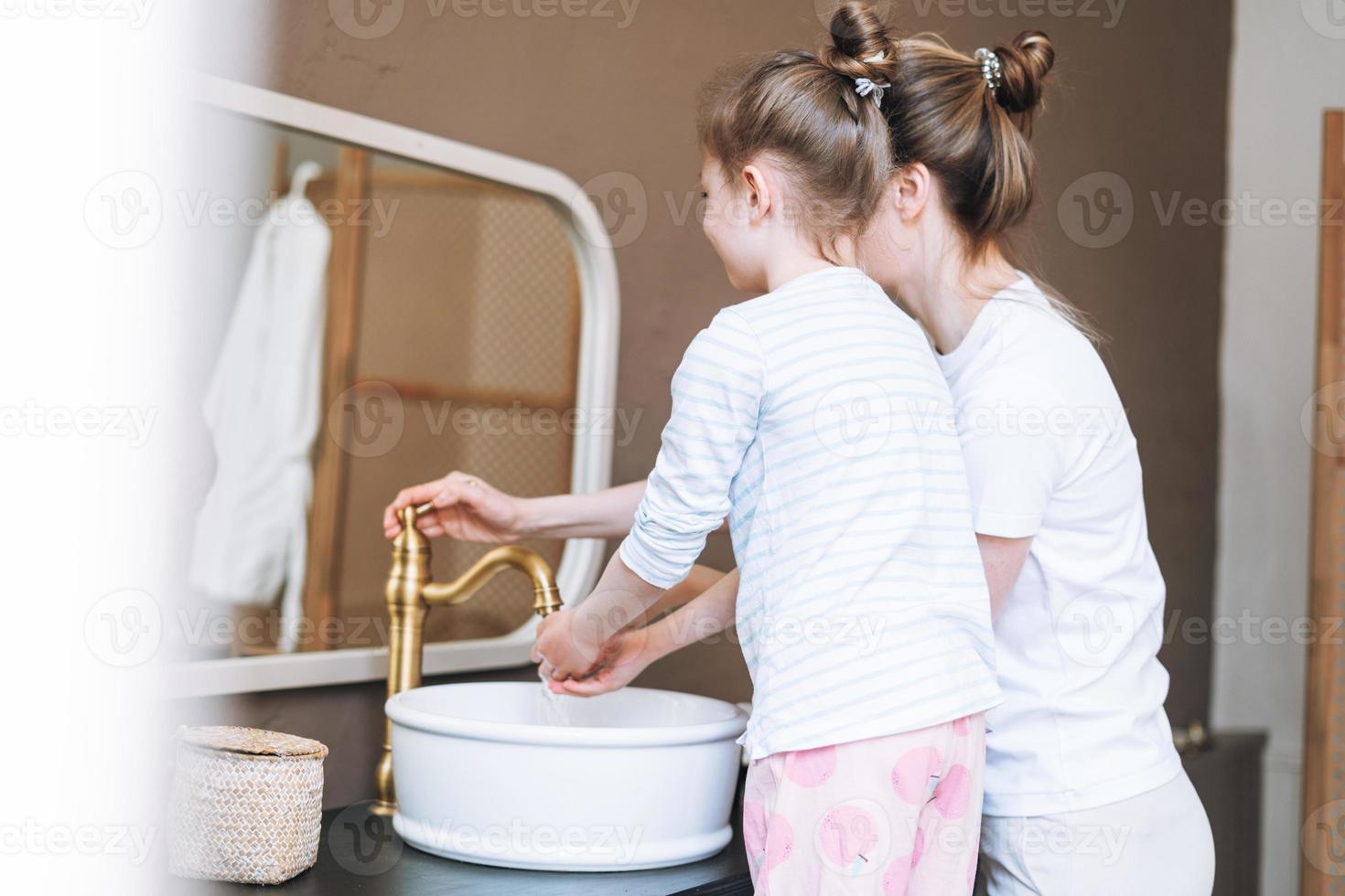 Young mother woman with long hair with little tween girl daughter in pajamas washing face and hands in the morning at home, family spending time together in bathroom photo