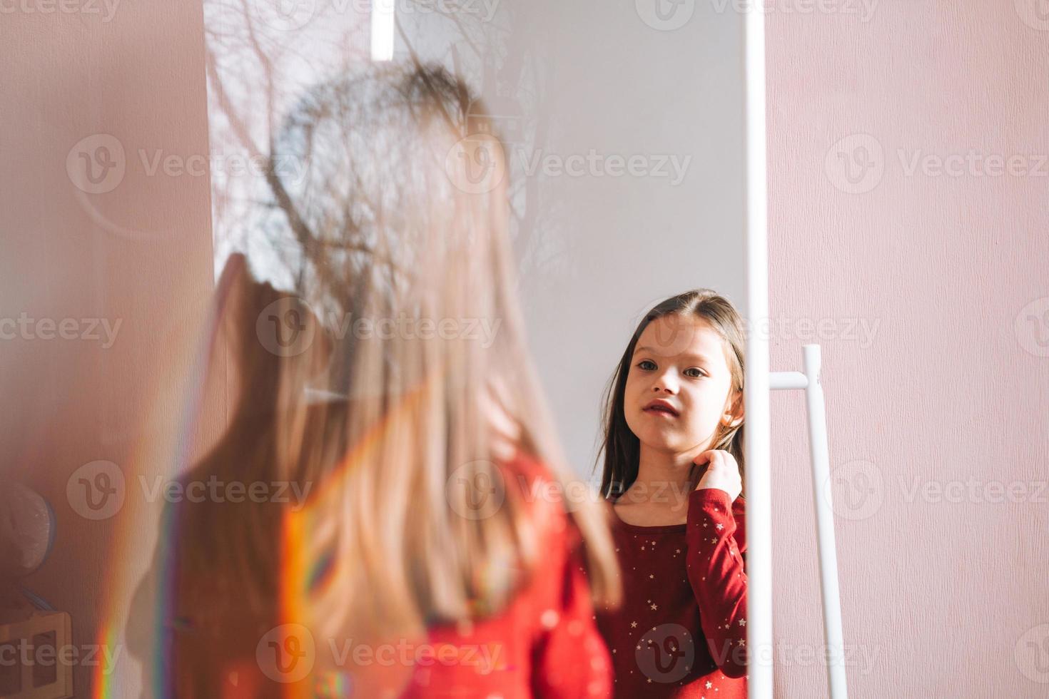 linda chica interpolada con el pelo largo y oscuro vestido rojo mirándose en el espejo en la habitación de los niños en la acogedora casa foto