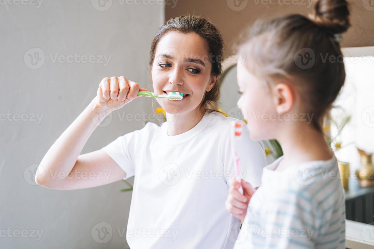 Young mother woman with long hair with little tween girl daughter in pajamas brushing their teeth in the morning at home photo