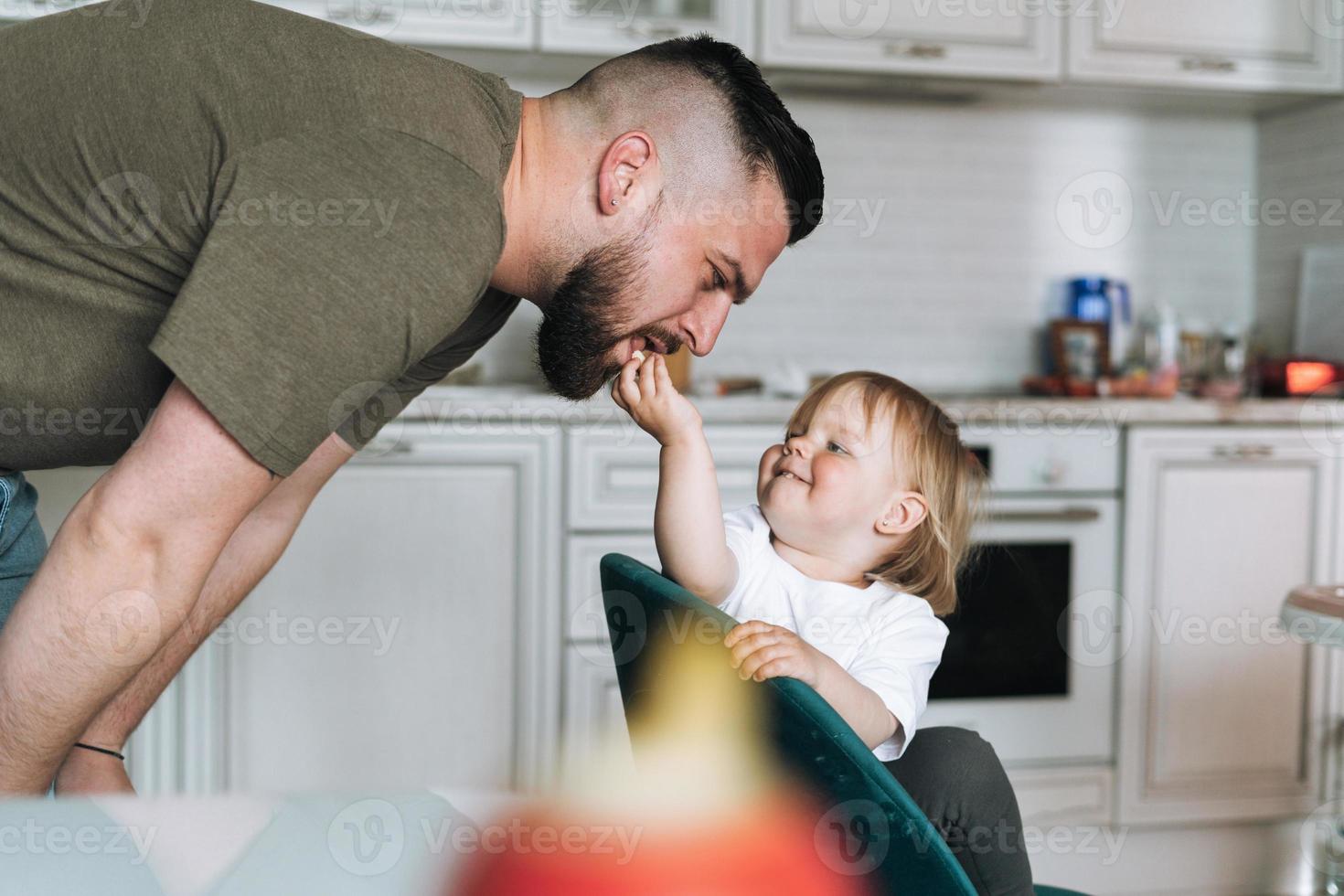 Happy father young man feeding and having fun with baby girl little daughter in kitchen at home photo