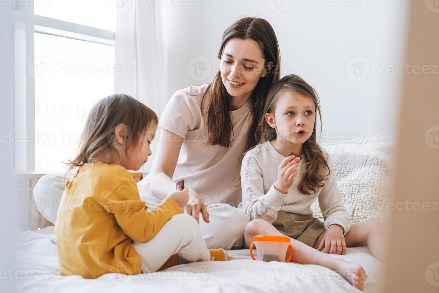 joven familia feliz con una madre madre con dos hijos niñas comiendo galletas en la habitación de la cama en casa foto