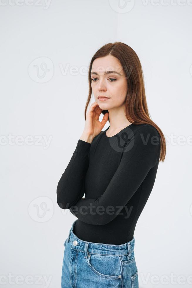 retrato vertical de una joven y hermosa mujer pensante con cabello largo oscuro en manga larga negra y jeans de longitud completa sobre fondo blanco aislado foto