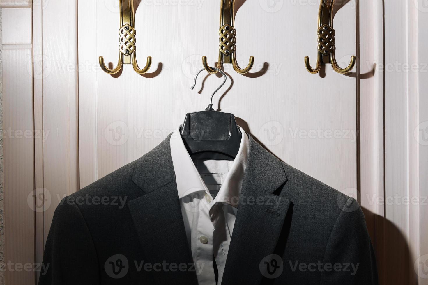 Man's dark grey suit jacket and white shirt hanging on gold metal hook on white wall background. photo