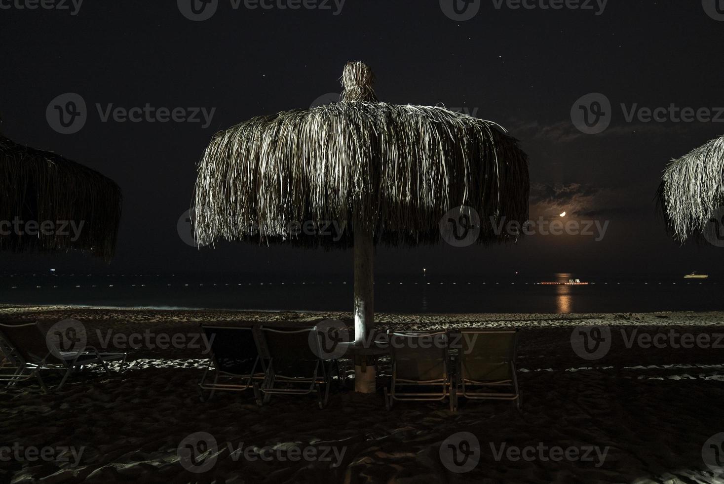 Empty deck chairs and thatched parasols arranged on sandy beach at night photo