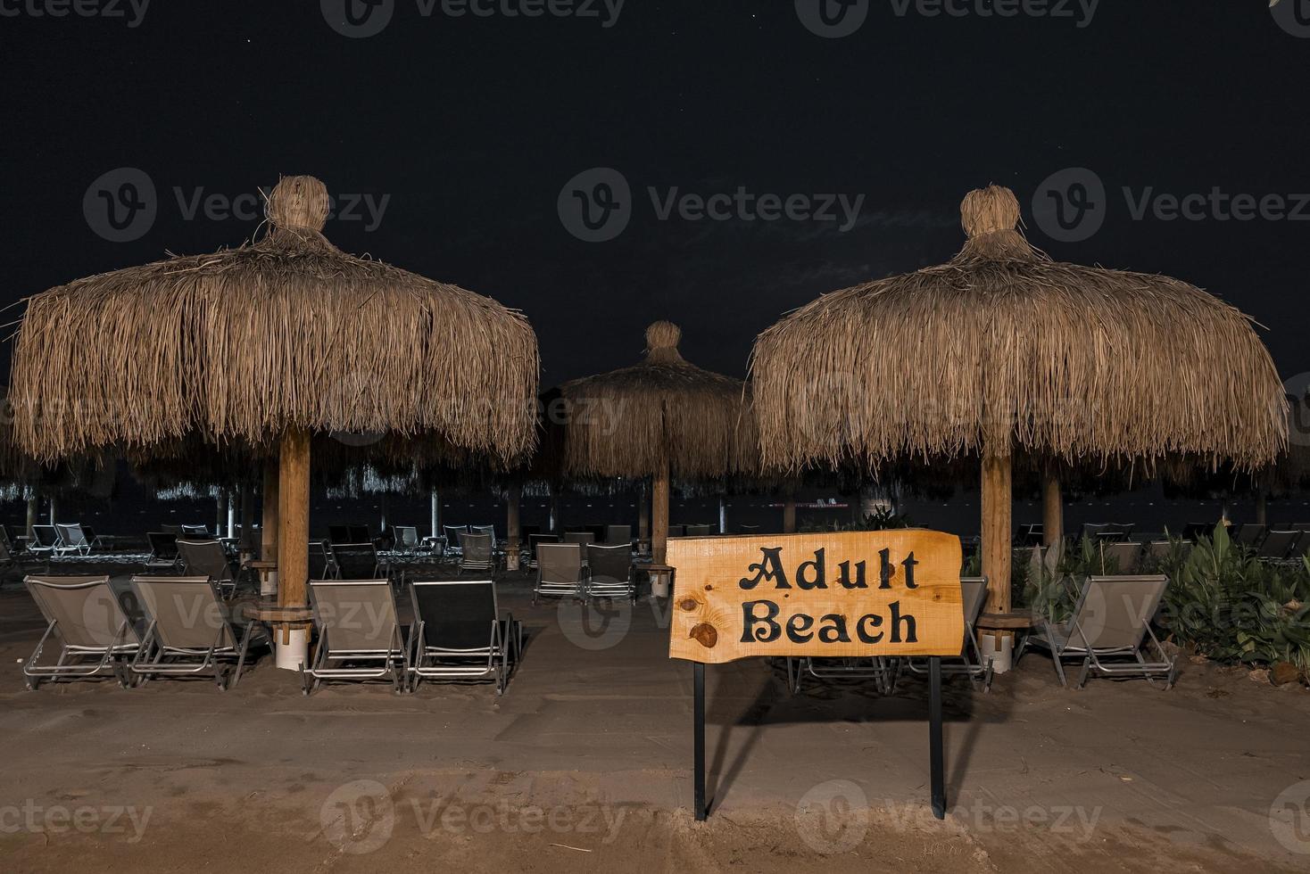 Adult beach text on signboard and deckchairs with parasols in background photo