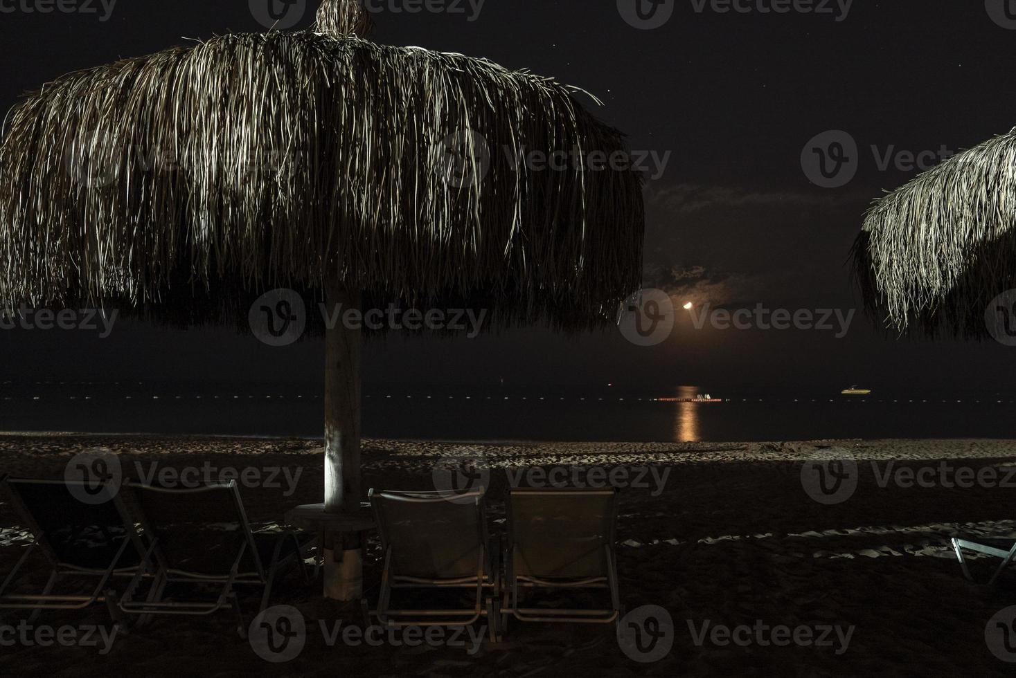 Empty deck chairs and thatched parasol arranged on sandy beach at night photo