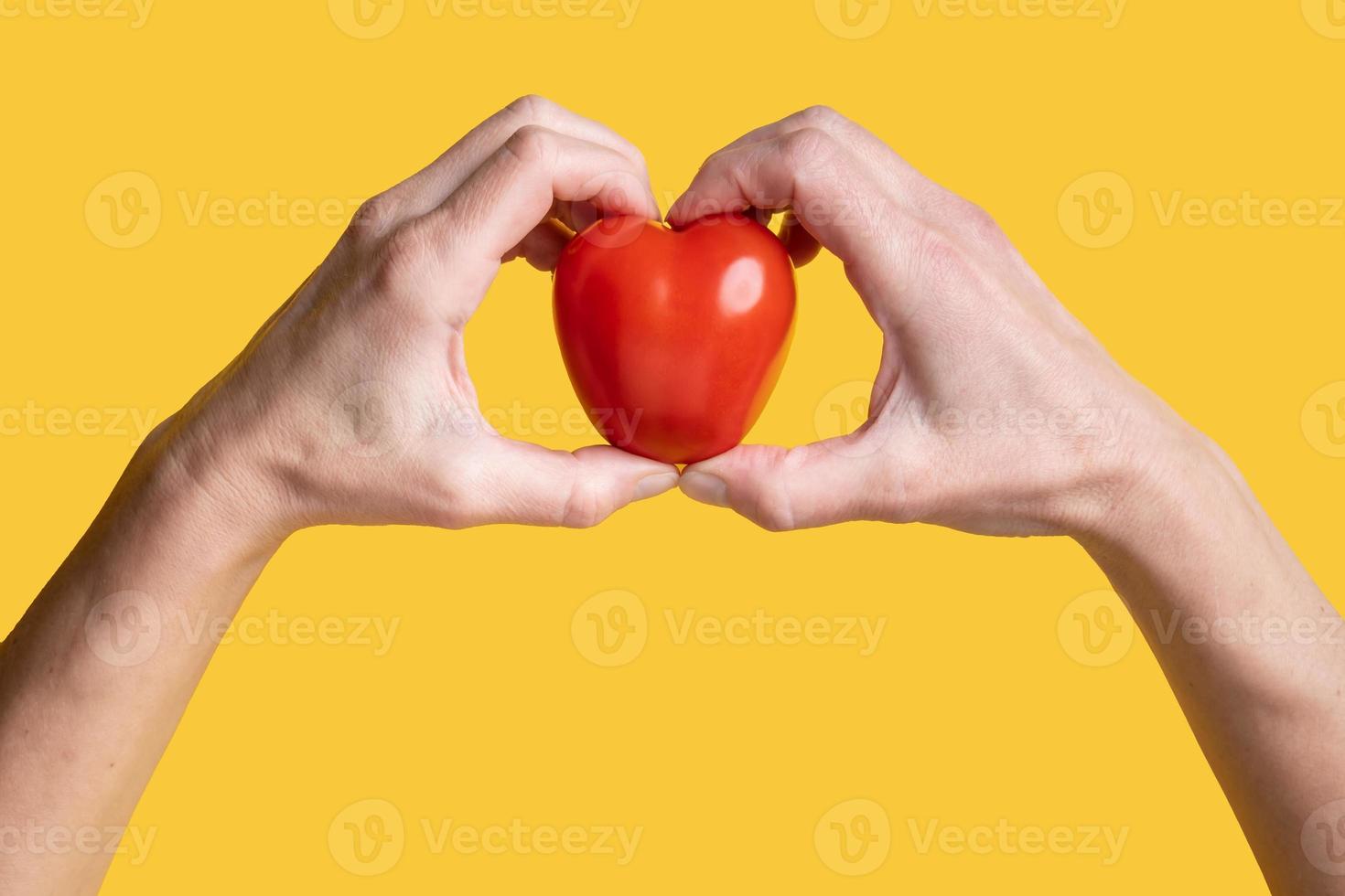 Red tomato in the form of a heart, a symbol of love in hands, on a yellow background. Healthy food concept. Valentine's Day. Top view. photo