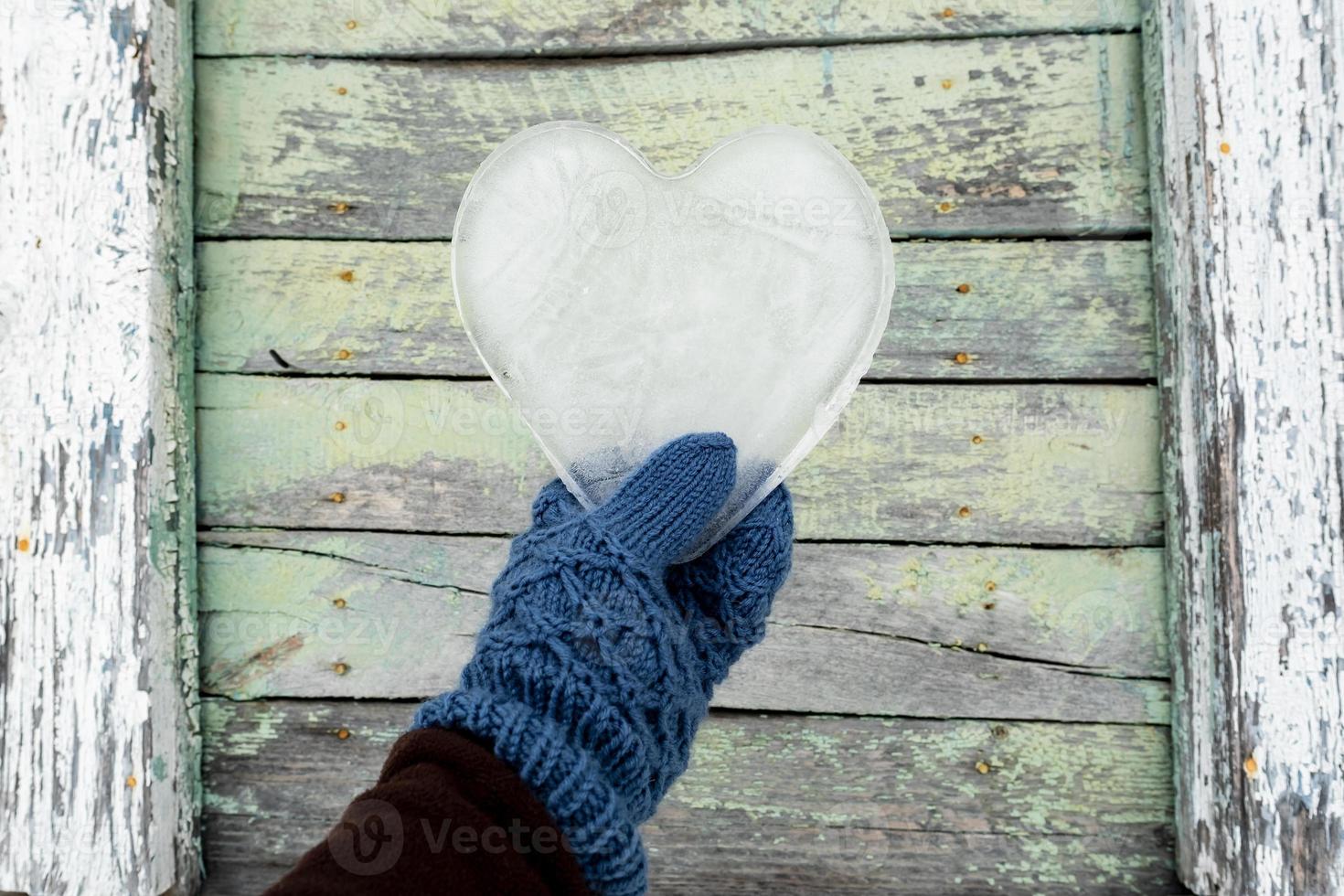 día de San Valentín. el corazón, un símbolo de amor, hecho de hielo, está sostenido por una mano en mitones contra el fondo de una vieja pared de madera. foto