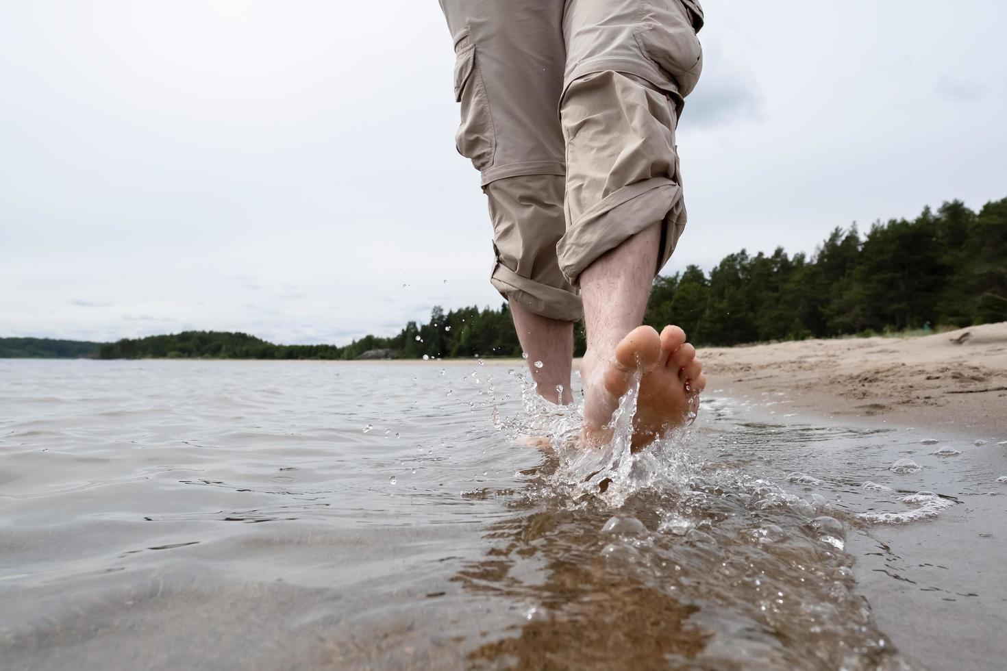 piernas masculinas descalzas con pantalones enrollados caminan sobre el agua a lo largo de la orilla arenosa contra el fondo del lago y los árboles. estilo de vida. foto