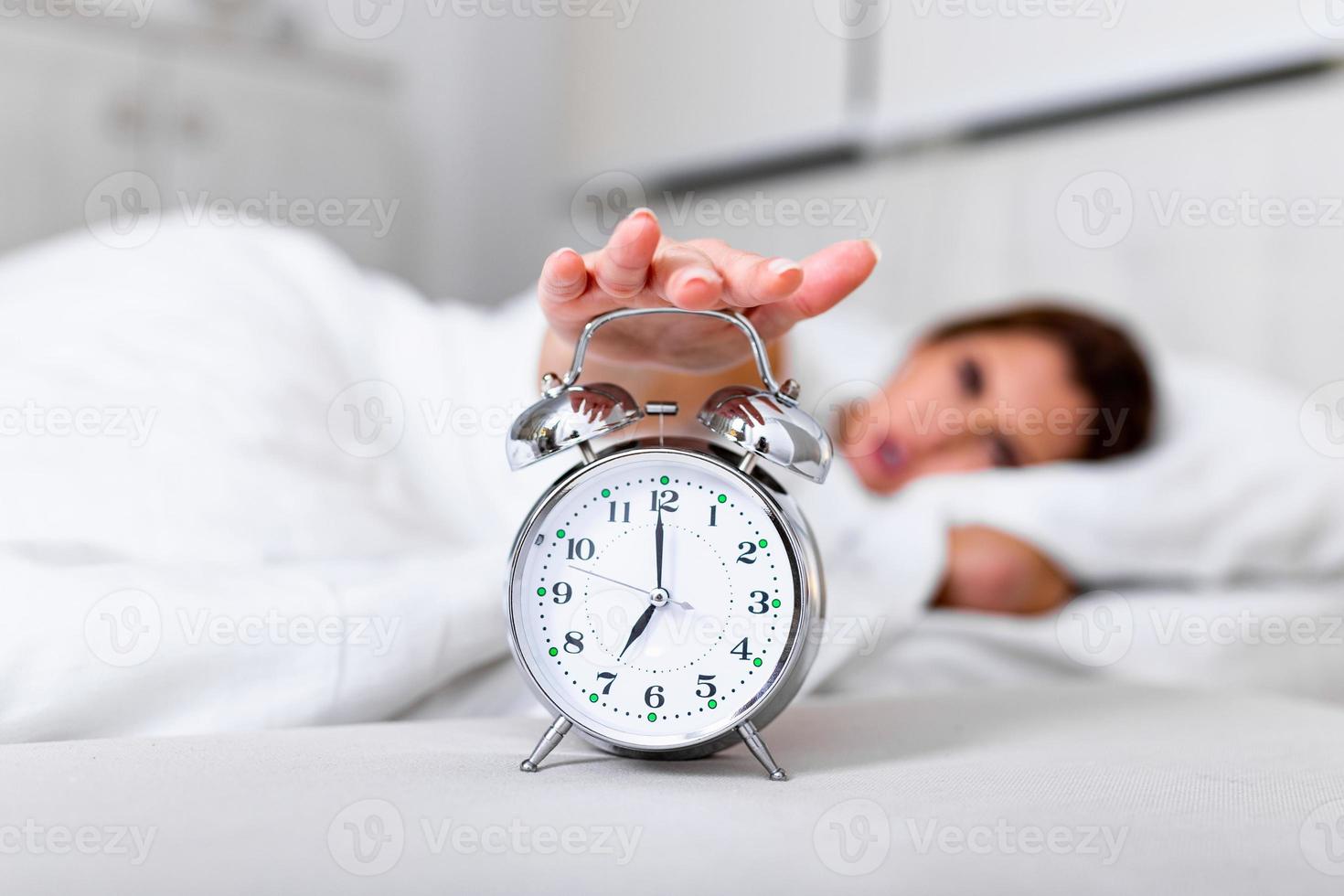 Woman lying in bed turning off an alarm clock in the morning at 7am. Hand turns off the alarm clock waking up at morning, girl turns off the alarm clock waking up in the morning from a call. photo