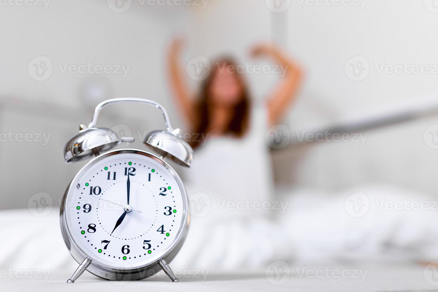 Closeup alarm clock having a good day with background happy woman stretching in bed after waking up, sunlight in morning. Waking up in the morning on time photo