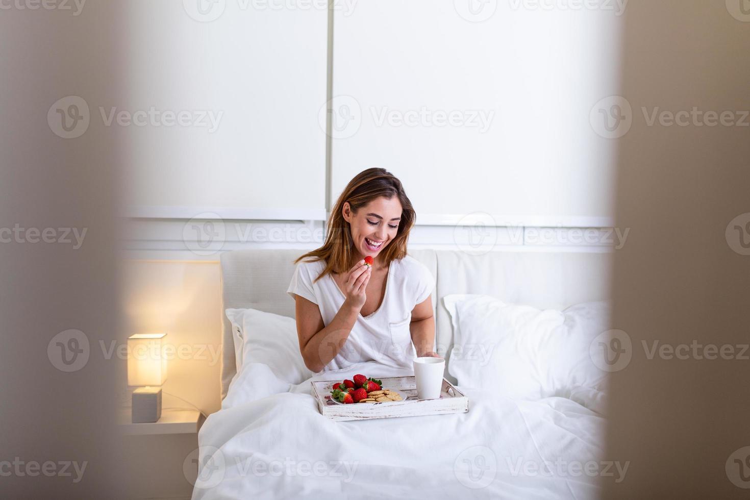 una mujer feliz y atractiva se despertó por la mañana en la cama. mujer desayunando en la habitación del hotel, mujer desayunando en la cama con frutas, café y galletas foto