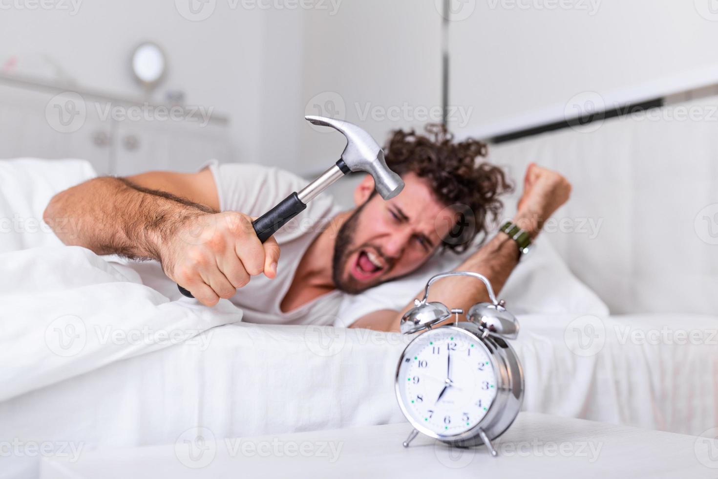 el joven intenta romper el despertador con un martillo, destruir el reloj. hombre acostado en la cama apagando un despertador con un martillo por la mañana a las 7 am. foto