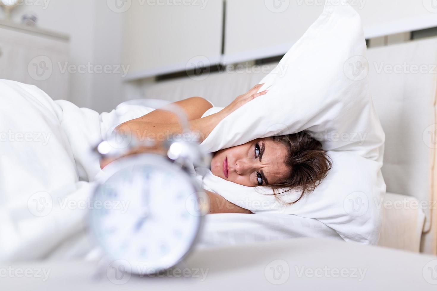 Frustrated woman covering her ears with her pillow from alarm clock in the morning. Portrait of annoyed young woman covering ears with pillow due to ringing alarm clock in morning, while lying in bed photo