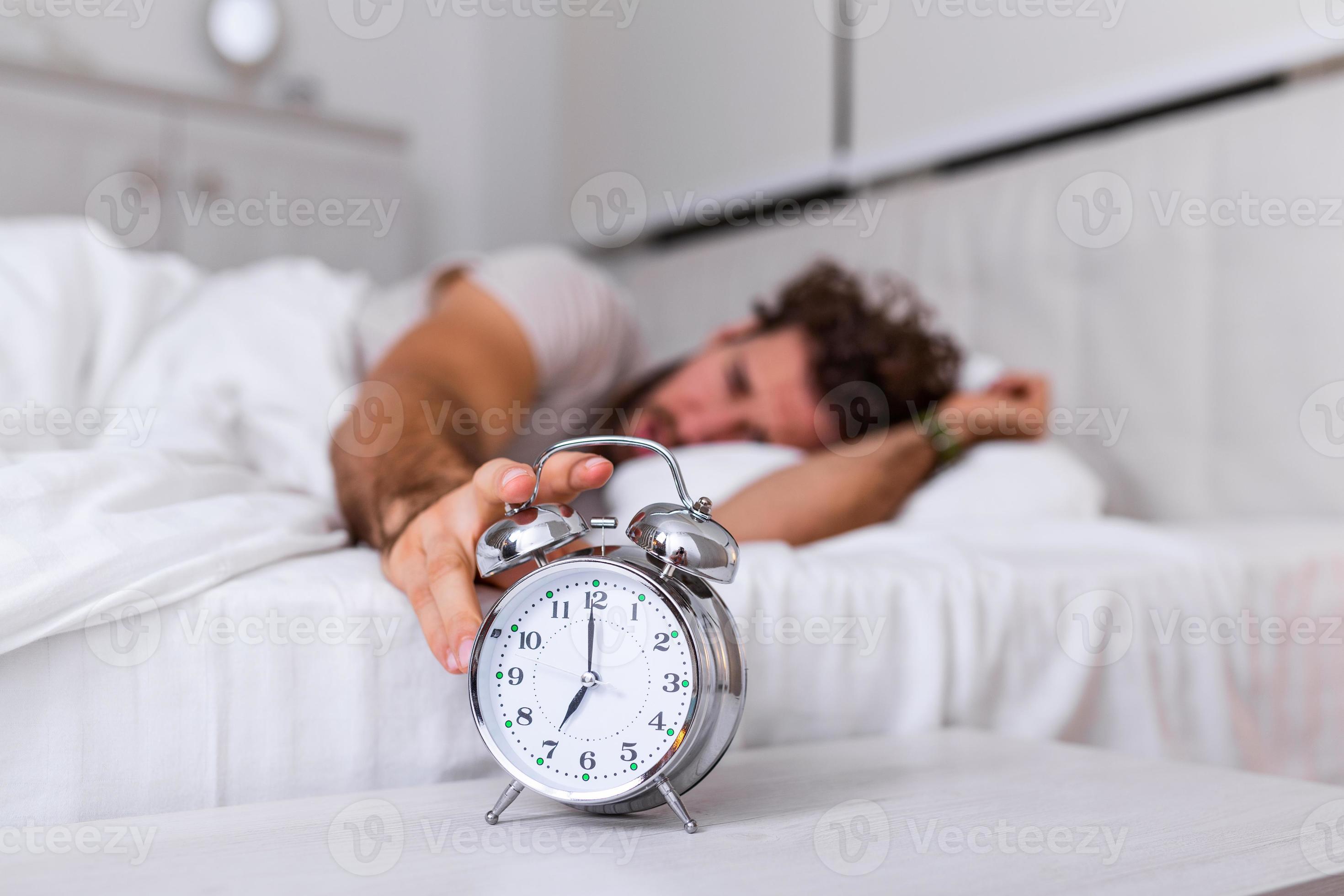 Man lying in bed turning off an alarm clock in the morning at 7am. Hand
