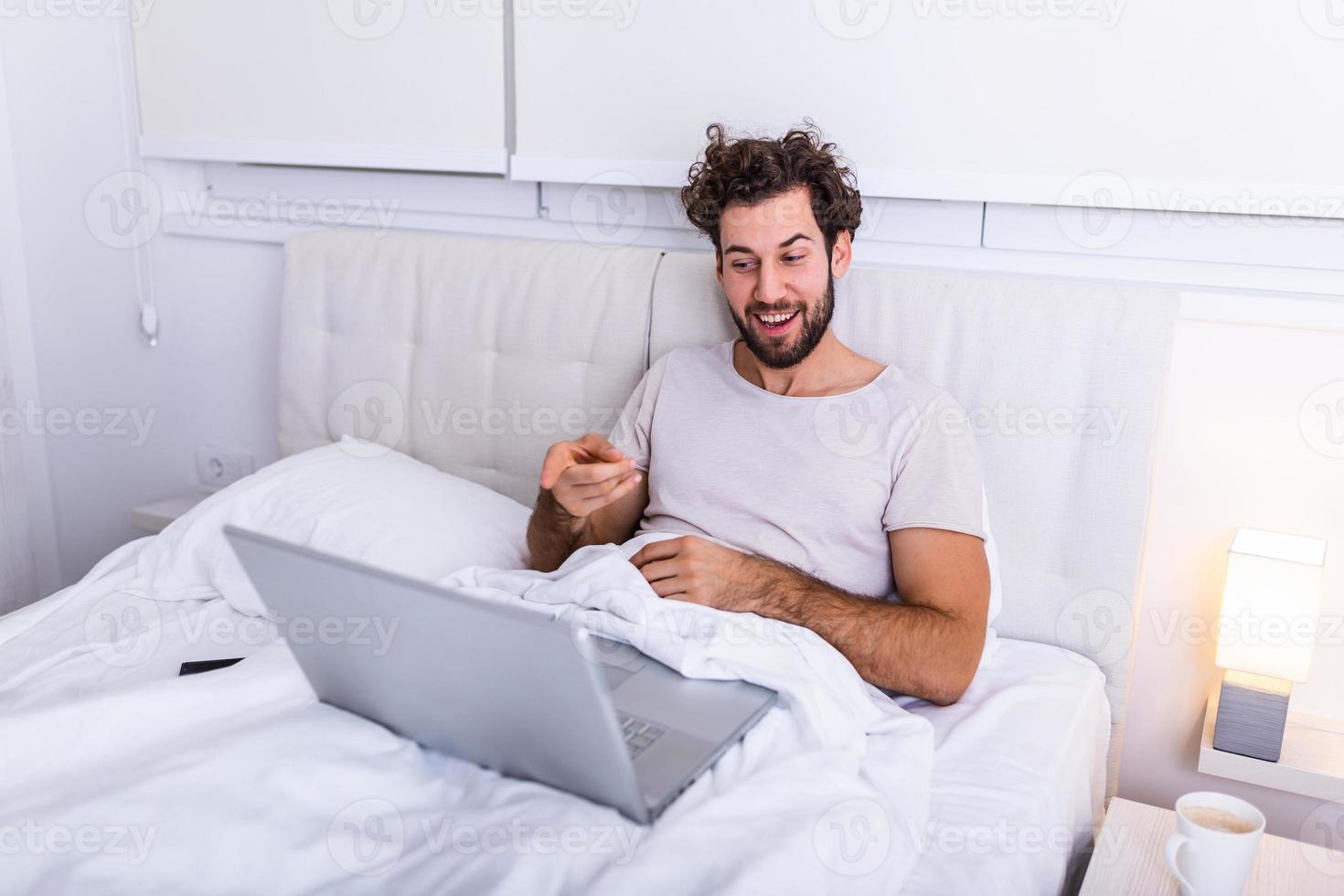 Young Caucasian man has morning coffee, sits on bed talking video call , reads book, prepares for examination, uses laptop computer for surfing internet, dressed in casual domestic clothes. photo