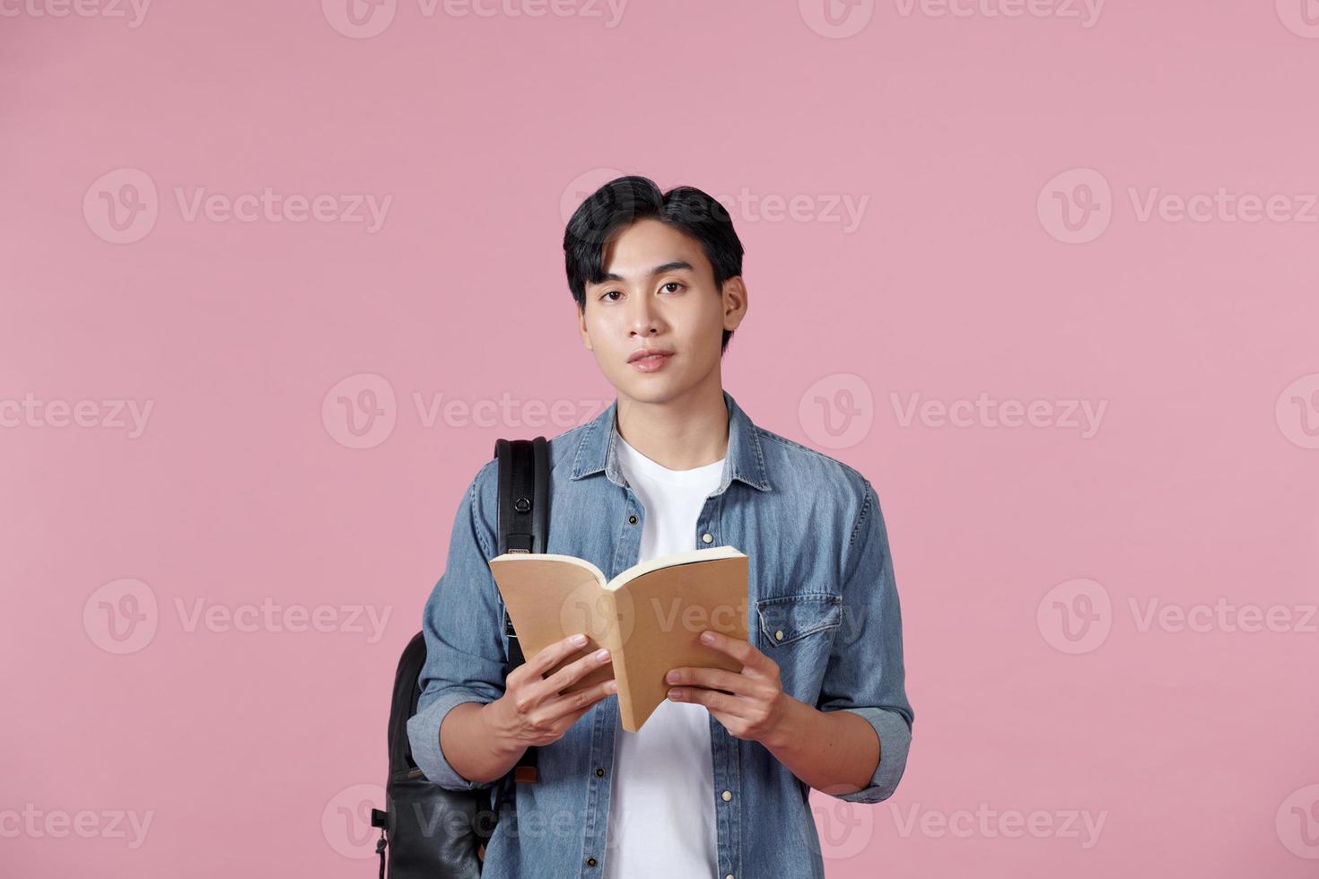 A young student carrying a backpack and holding an open book reading, isolated on a pink background. photo