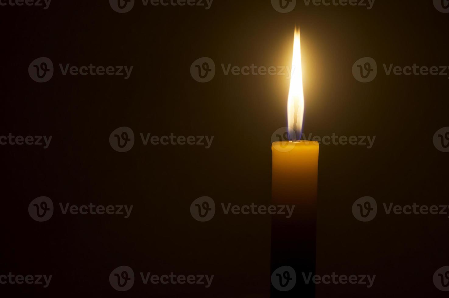 A single burning candle flame or light glowing on a yellow candle isolated on red or dark background on table in church for Christmas, funeral or memorial service photo