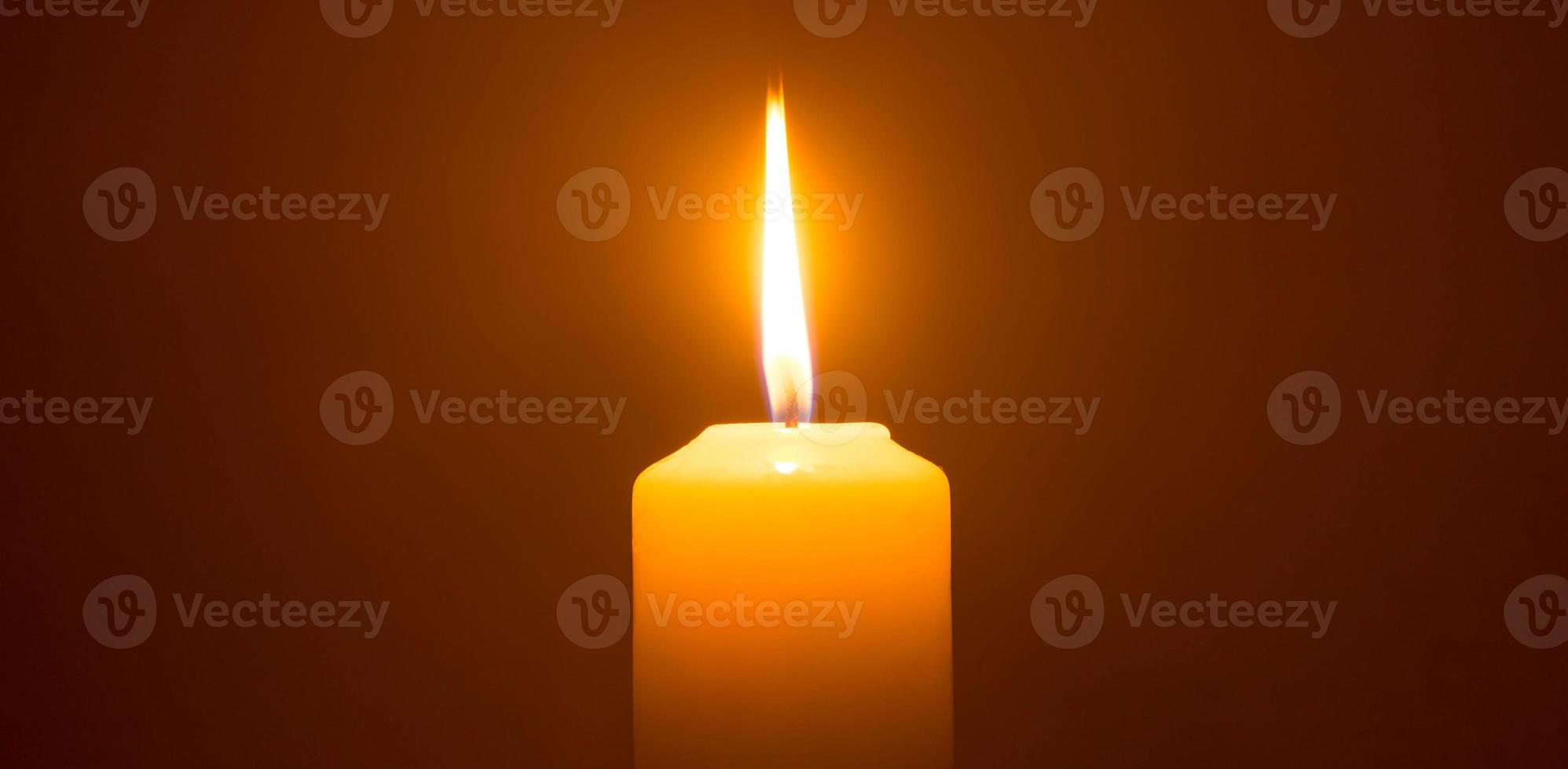 A single burning candle flame or light glowing on a big yellow candle isolated on red or dark background on table in church for Christmas, funeral or memorial service photo