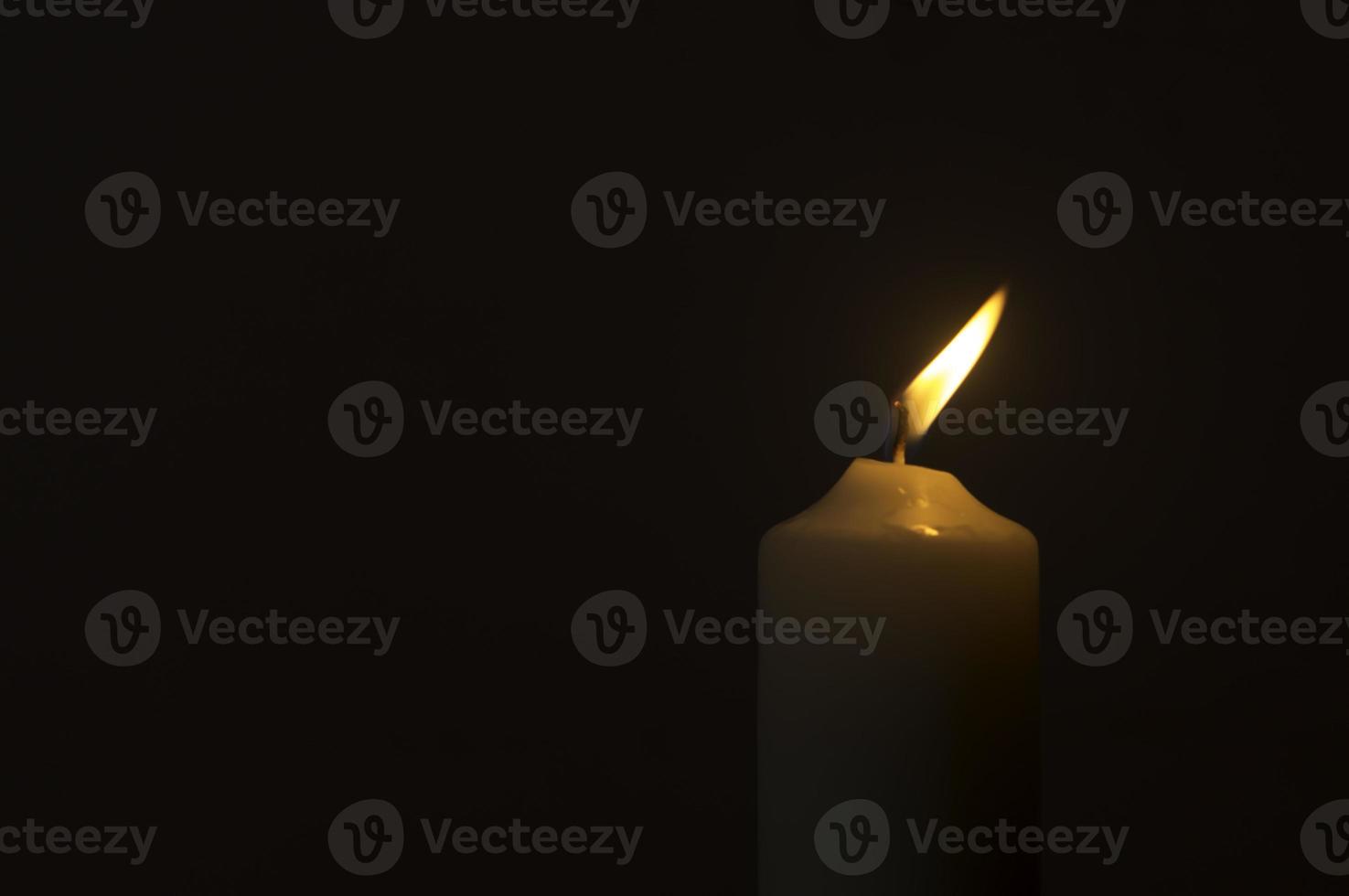 A single burning candle flame or light glowing on a white candle on black or dark background on table in church for Christmas, funeral or memorial service photo