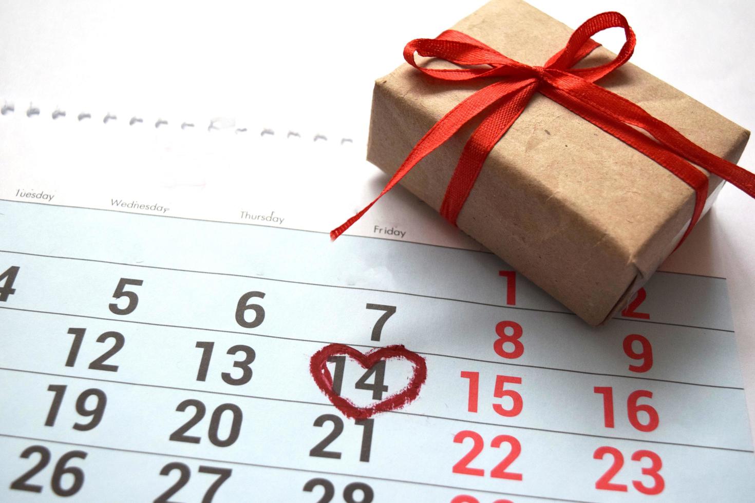 Calendar page with heart written in red on February 14 of Valentine's Day, gift box on white background. Top view with space for greetings. Concept of Valentine's day, holiday photo