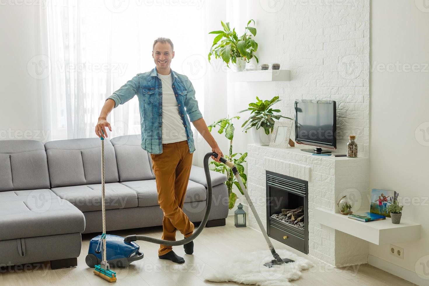 Caucasian handsome serious young man vacuuming cleaning modern apartment, clean up, housekeeper, guy doing domestic tasks and cleans living room using vacuum cleaner, householding photo