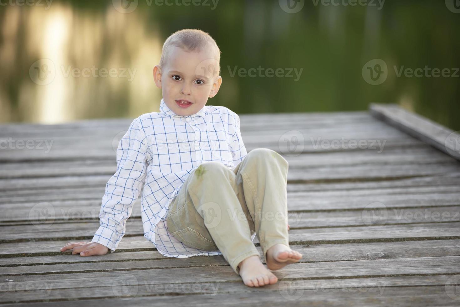 A handsome little boy sits on a wooden bridge by the lake and looks at the camera. photo