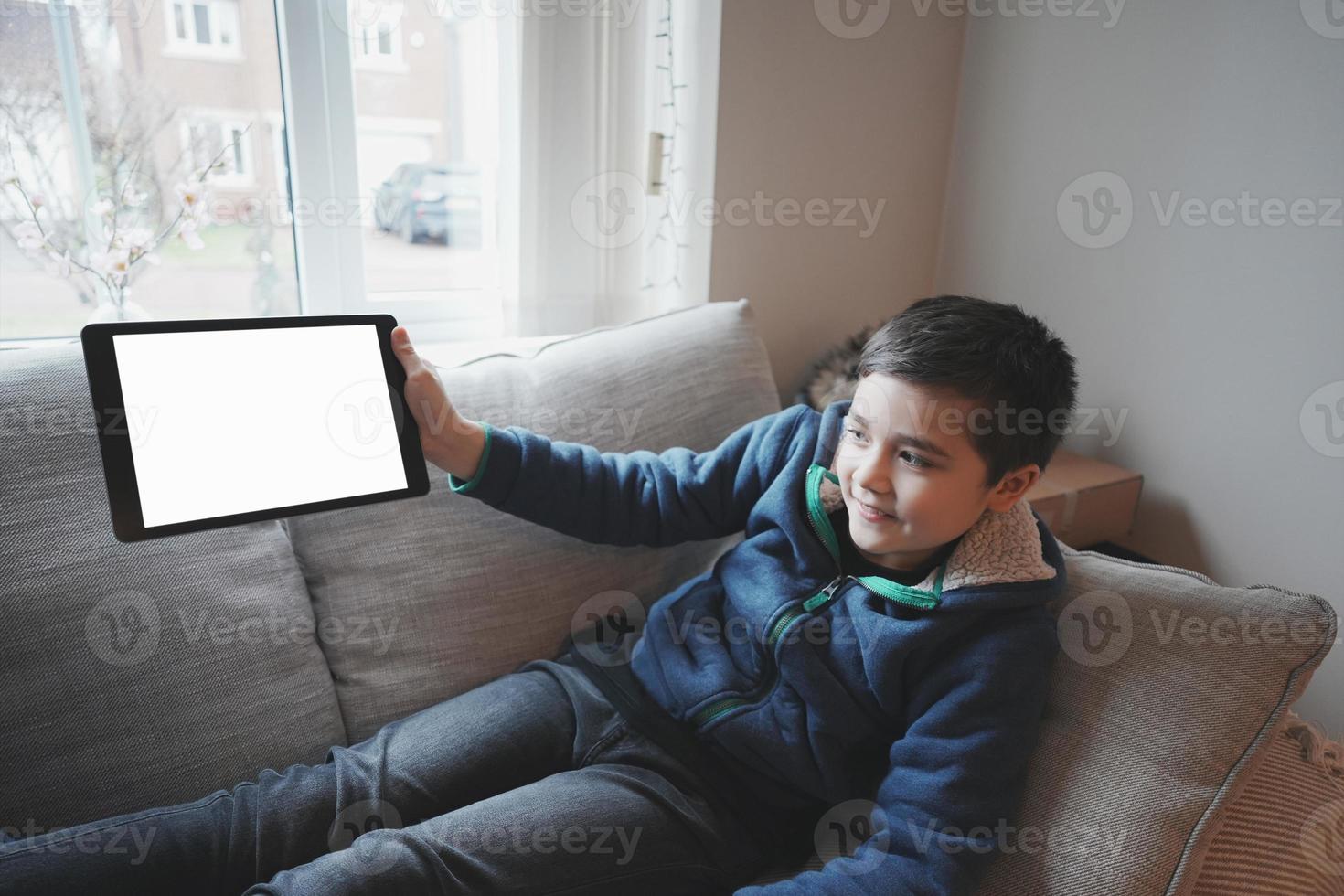 Happy Child with tablet computer. Portrait Kid Holding And Showing Digital Tablet With Empty Screen  Mockup, Young boy sitting on sofa relaxing and playing with touch pad at home photo