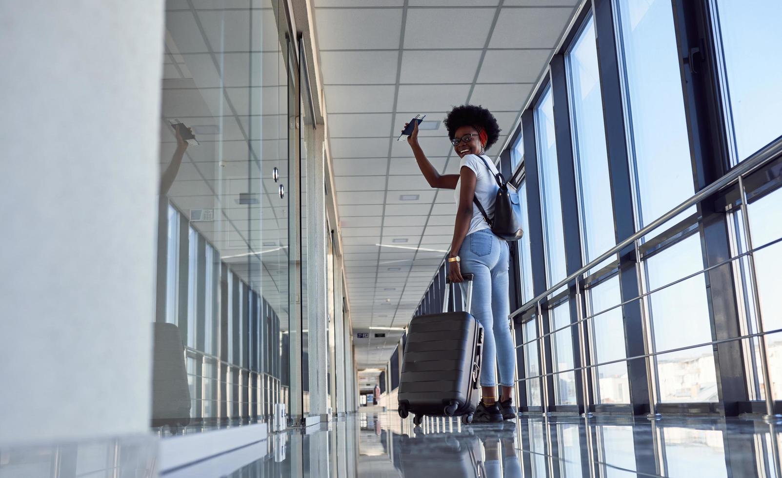 vista trasera si una joven pasajera afroamericana con ropa informal que está en el aeropuerto con equipaje foto