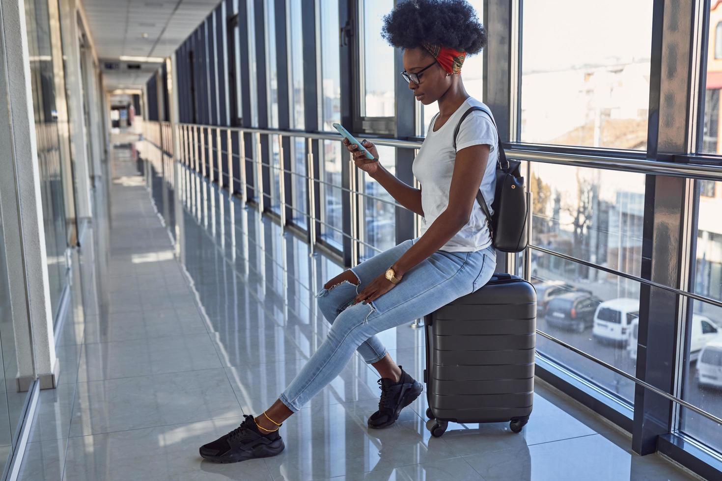 Young african american female passanger in casual clothes is in airport with baggage. Using phone photo