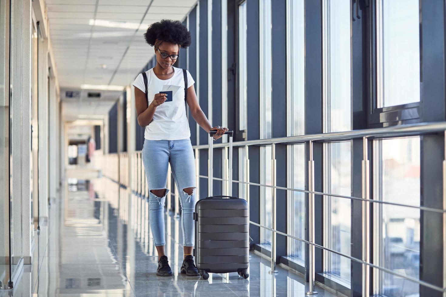 Young african american female passanger in casual clothes is in airport with baggage. Using phone photo